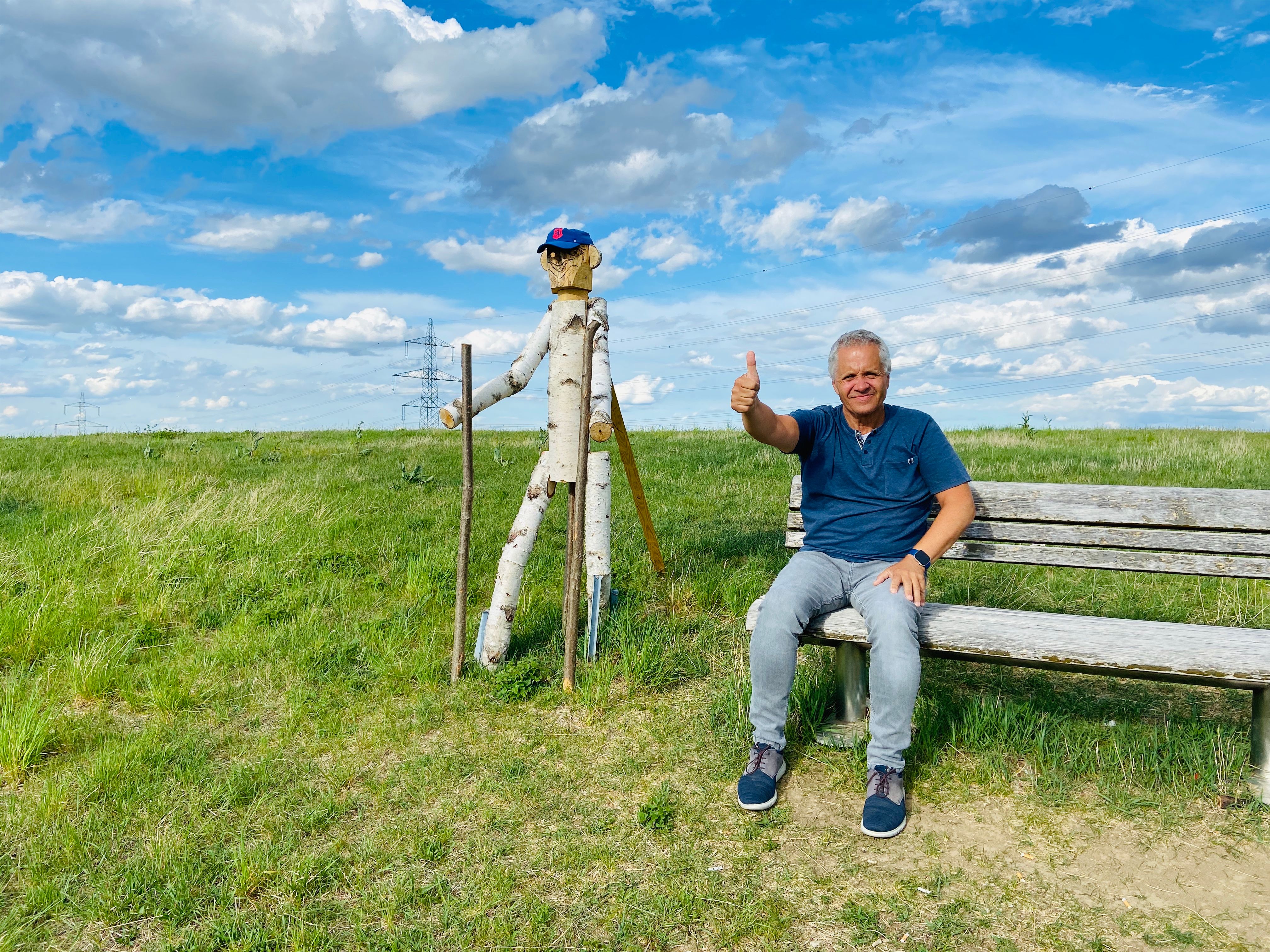 Ein Mann sitzt auf einer Bank neben einer Holzfigur auf einem Feld unter blauem Himmel.