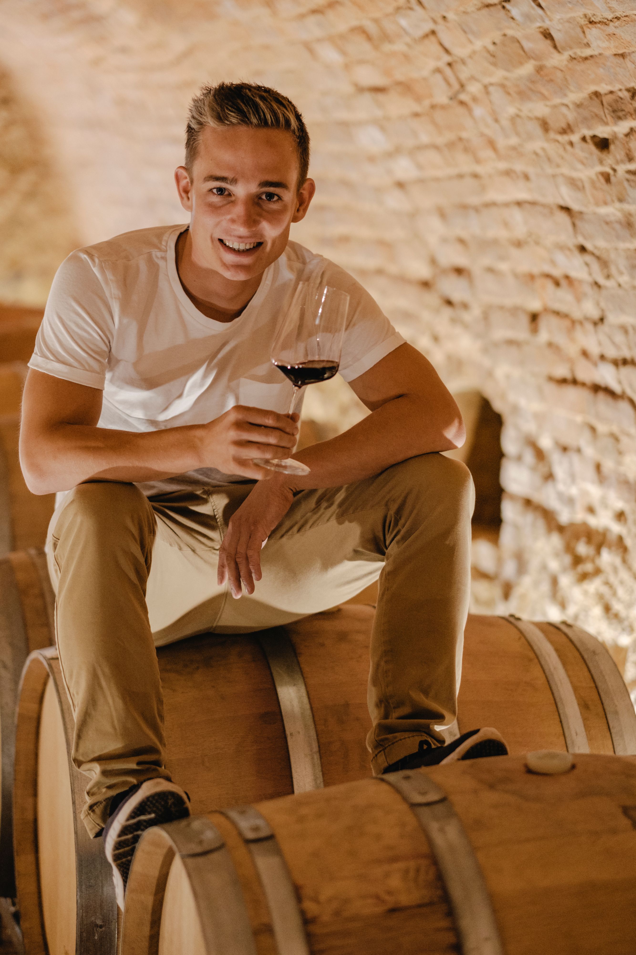 A young man sits on a wine barrel in a cellar and holds a glass of red wine in his hand.