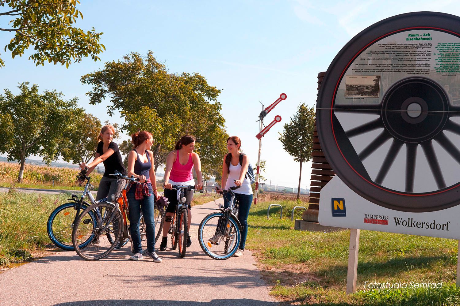 Vier Frauen mit Fahrrädern auf einem Radweg neben einem großen Rad-Denkmal in Wolkersdorf.