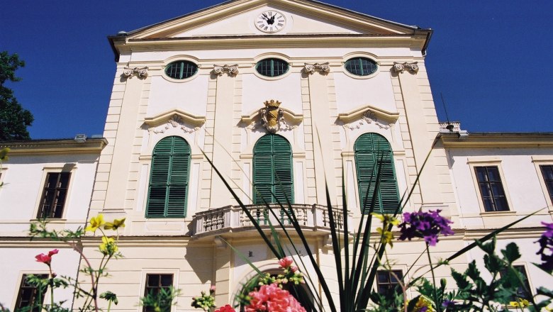 Facade of Kirchstetten Castle with green shutters and clock, surrounded by colorful flowers.