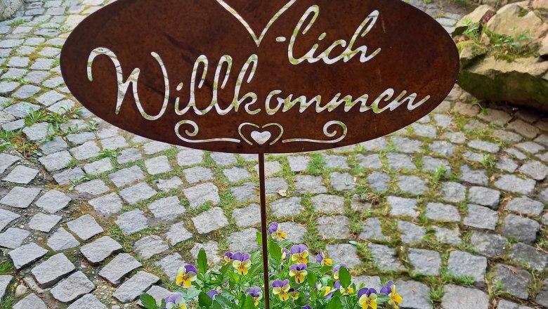 Metallschild mit 'Herzlich Willkommen' über einem Topf mit gelb-violetten Blumen auf einem gepflasterten Weg.