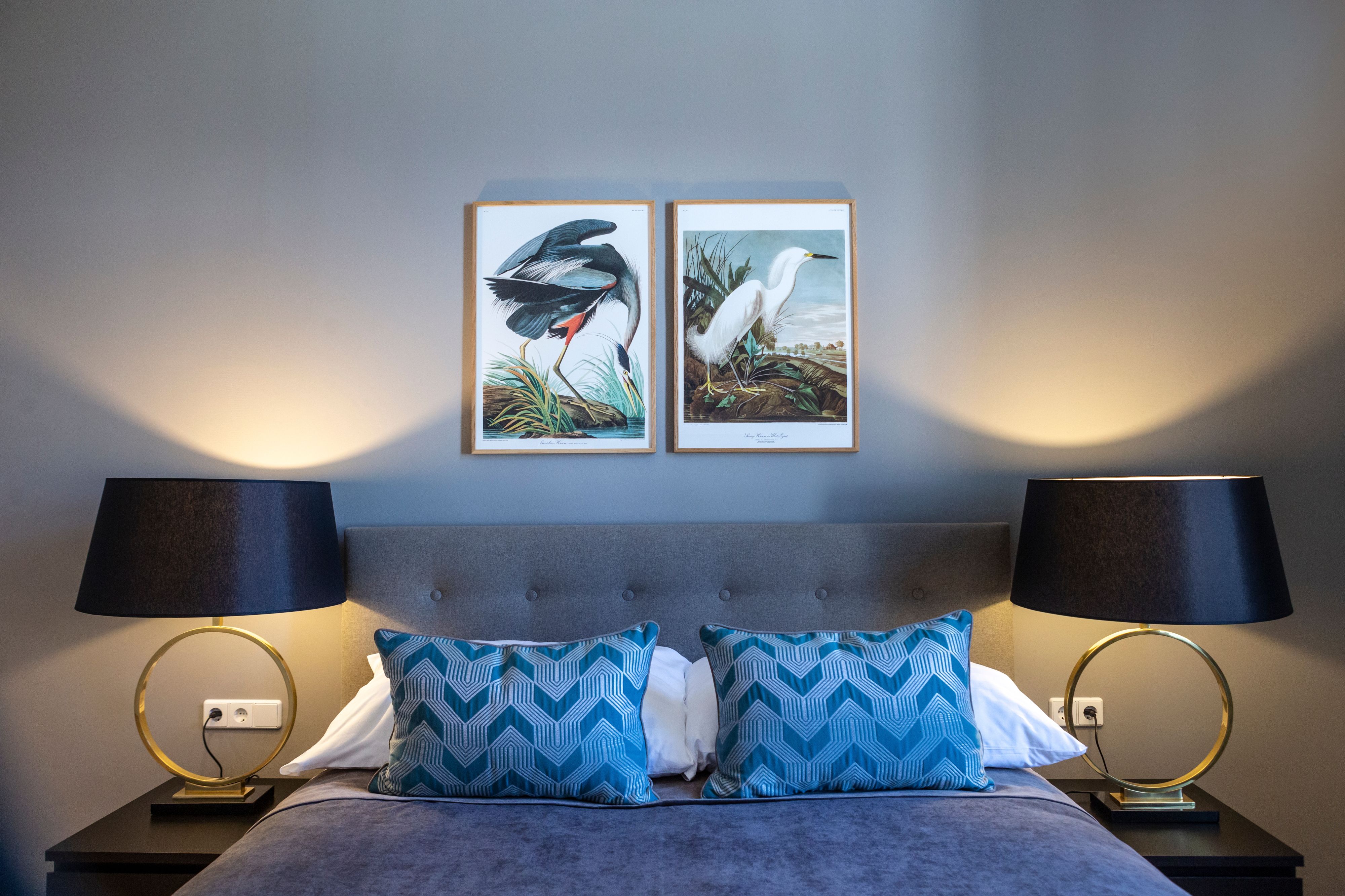 Doppelzimmer mit grauem Bett, blauen Kissen, zwei Nachttischlampen und Vogelbildern an der Wand.