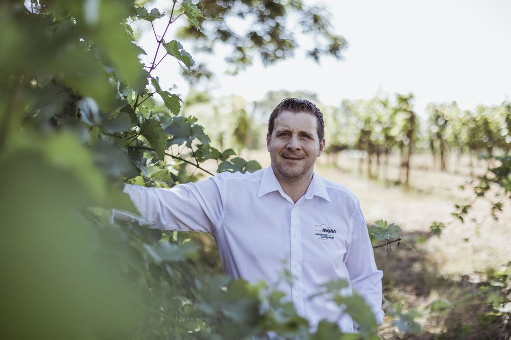 Mann in weißem Hemd steht in einem Weinberg, umgeben von Reben.