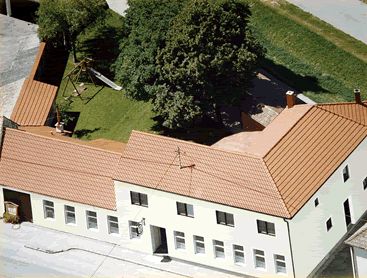 Letecký pohled na komplex budov s červenou střechou a vnitřním dvorem se stromy a dětským hřištěm.