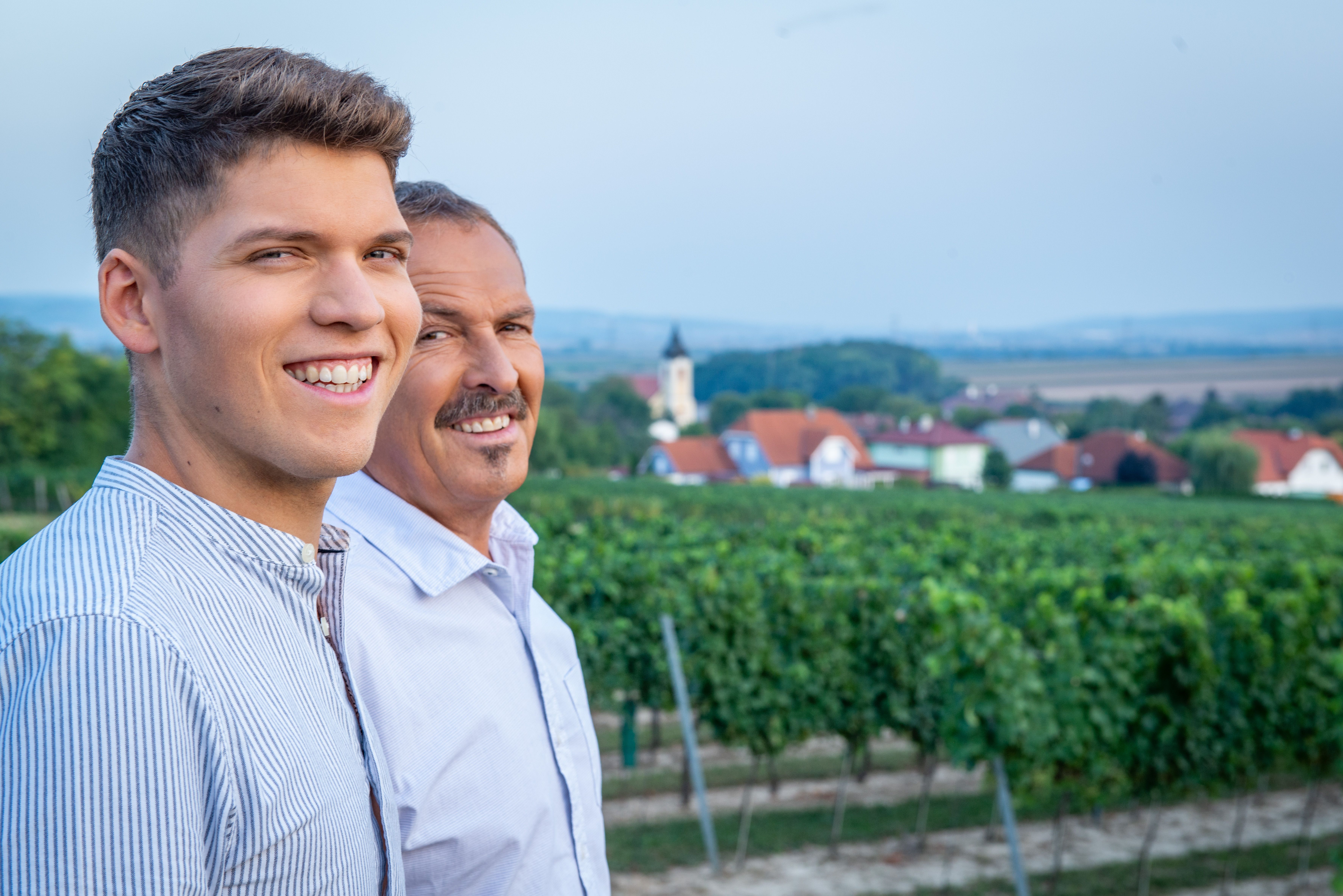 Two men smile in a vineyard with a village in the background.