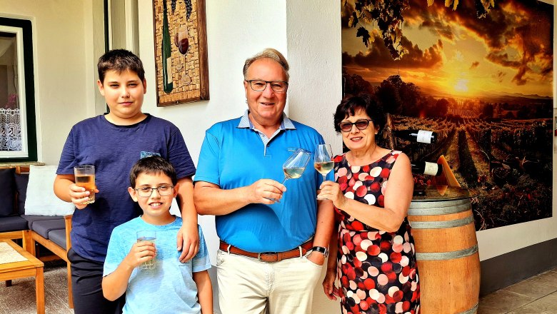 Eine Familie steht vor einem Weinfass und einem Bild von Weinbergen, hält Gläser und lächelt in die Kamera.