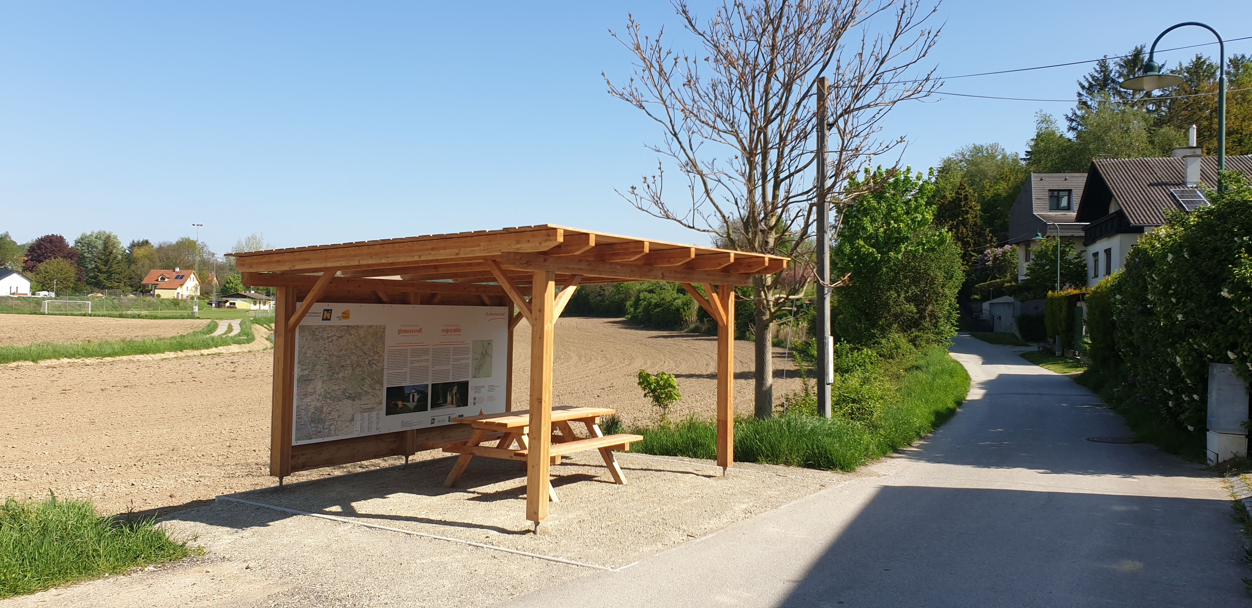 A rest area with a wooden roof and a bench next to a dirt road in a rural setting.