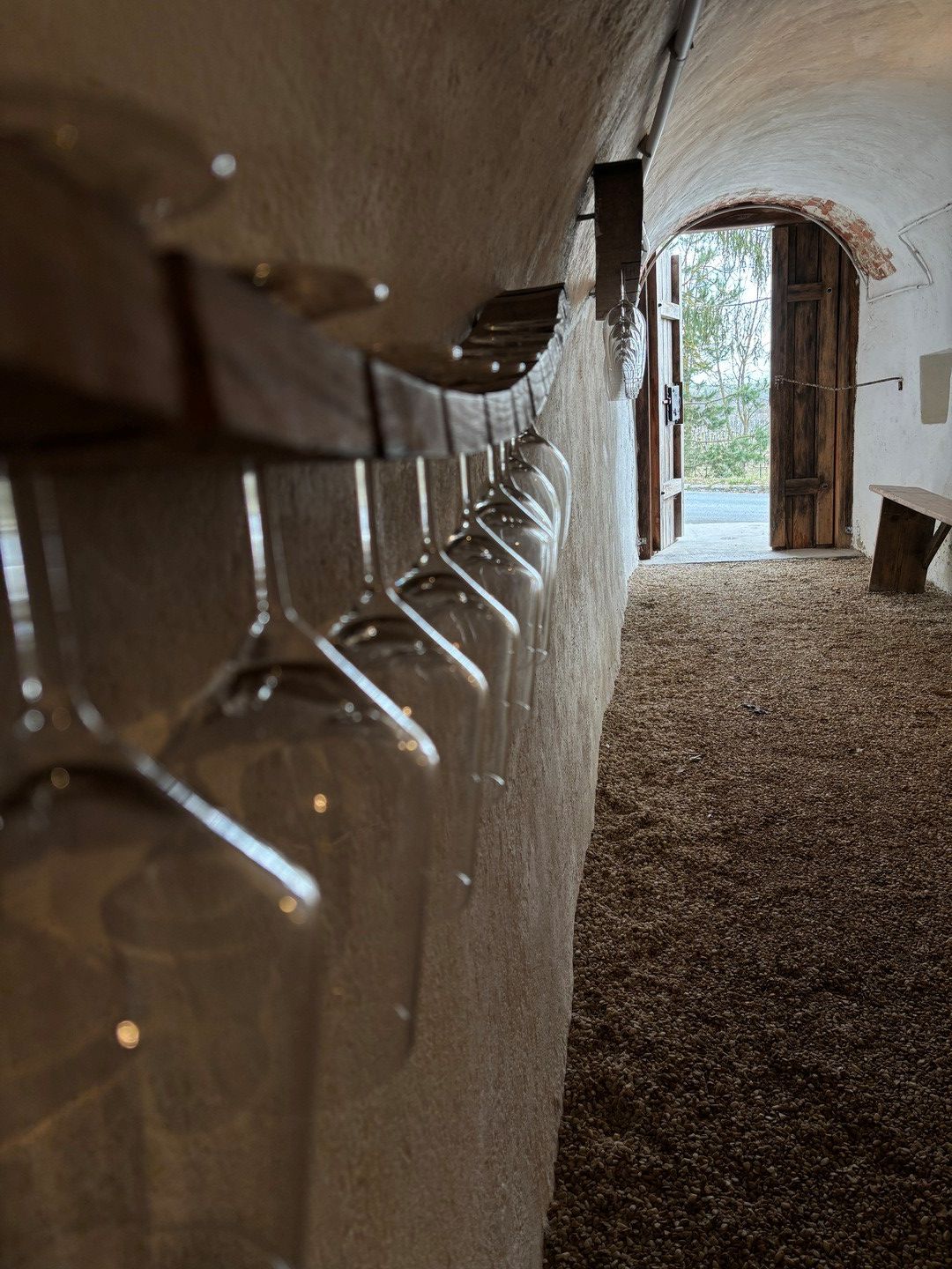 Blick in die Trinkstation Weinbau Ackerl mit hängenden Weingläsern an einer Wand, Gewölbe und offenem Holztor nach draußen.