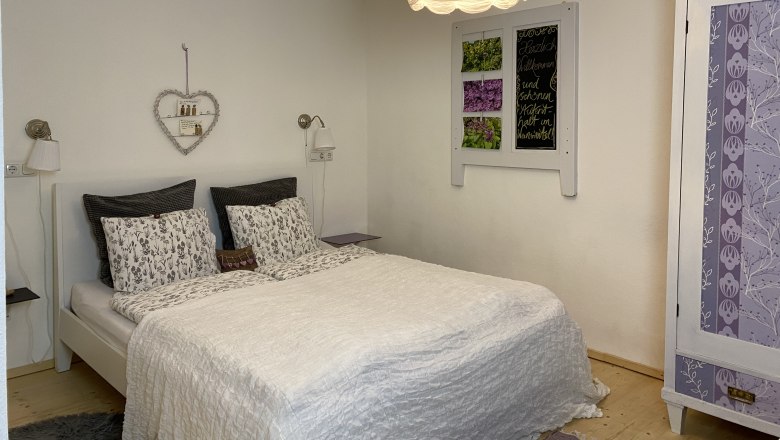 Gemütliches Doppelzimmer mit weißem Bett, dekorativem Herzregal und lila Schrank.