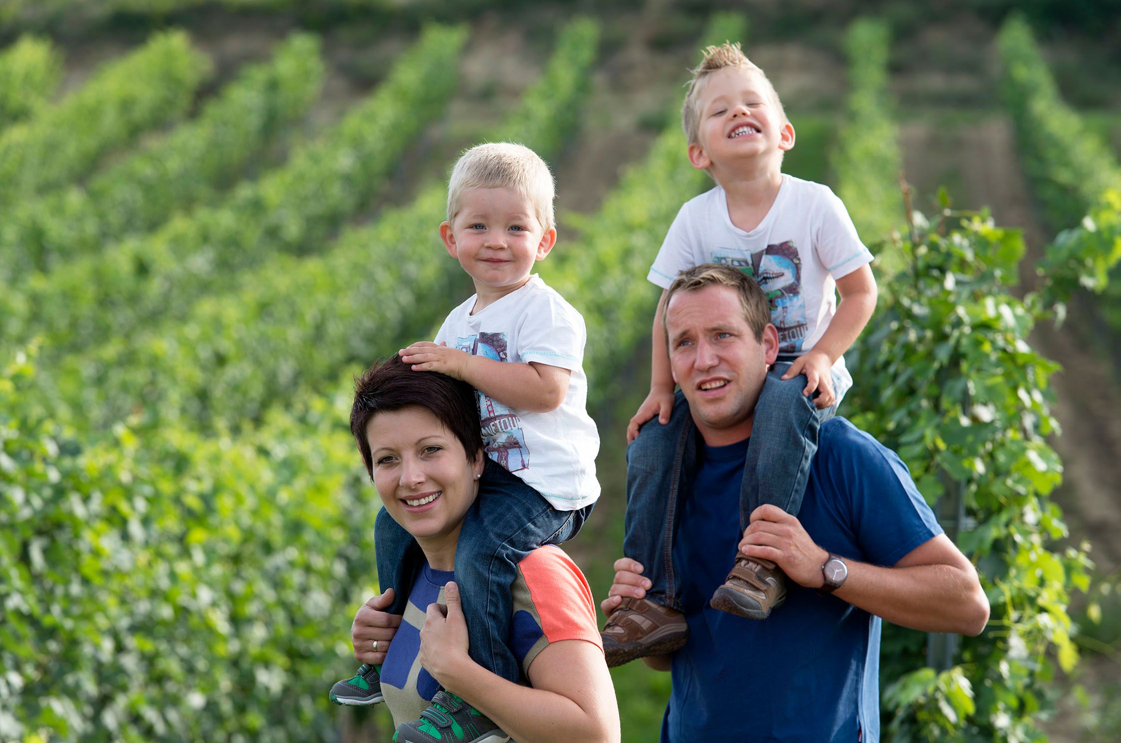 Family in a vineyard, two children on their parents' shoulders.
