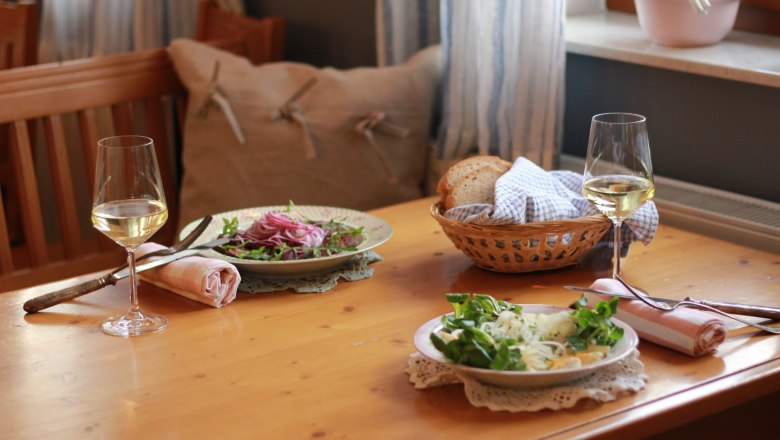 Two plates of salad and two glasses of white wine on a wooden table, with a bread basket next to them.