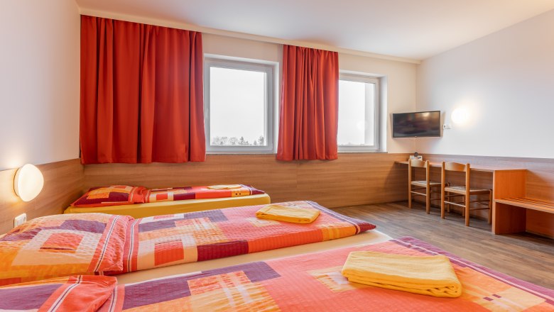 Hotelzimmer mit drei Betten, roten Vorhängen und einem Fernseher an der Wand.