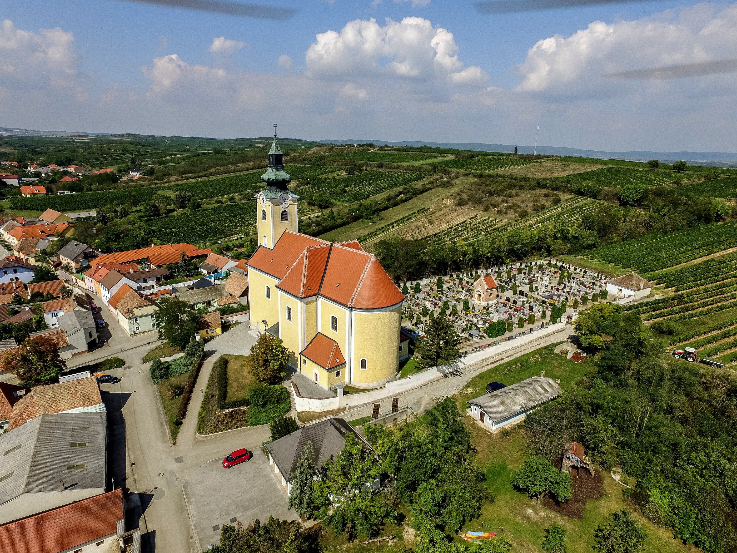 Luftaufnahme der Pfarrkirche in Röschitz mit umliegendem Friedhof und Weinbergen.