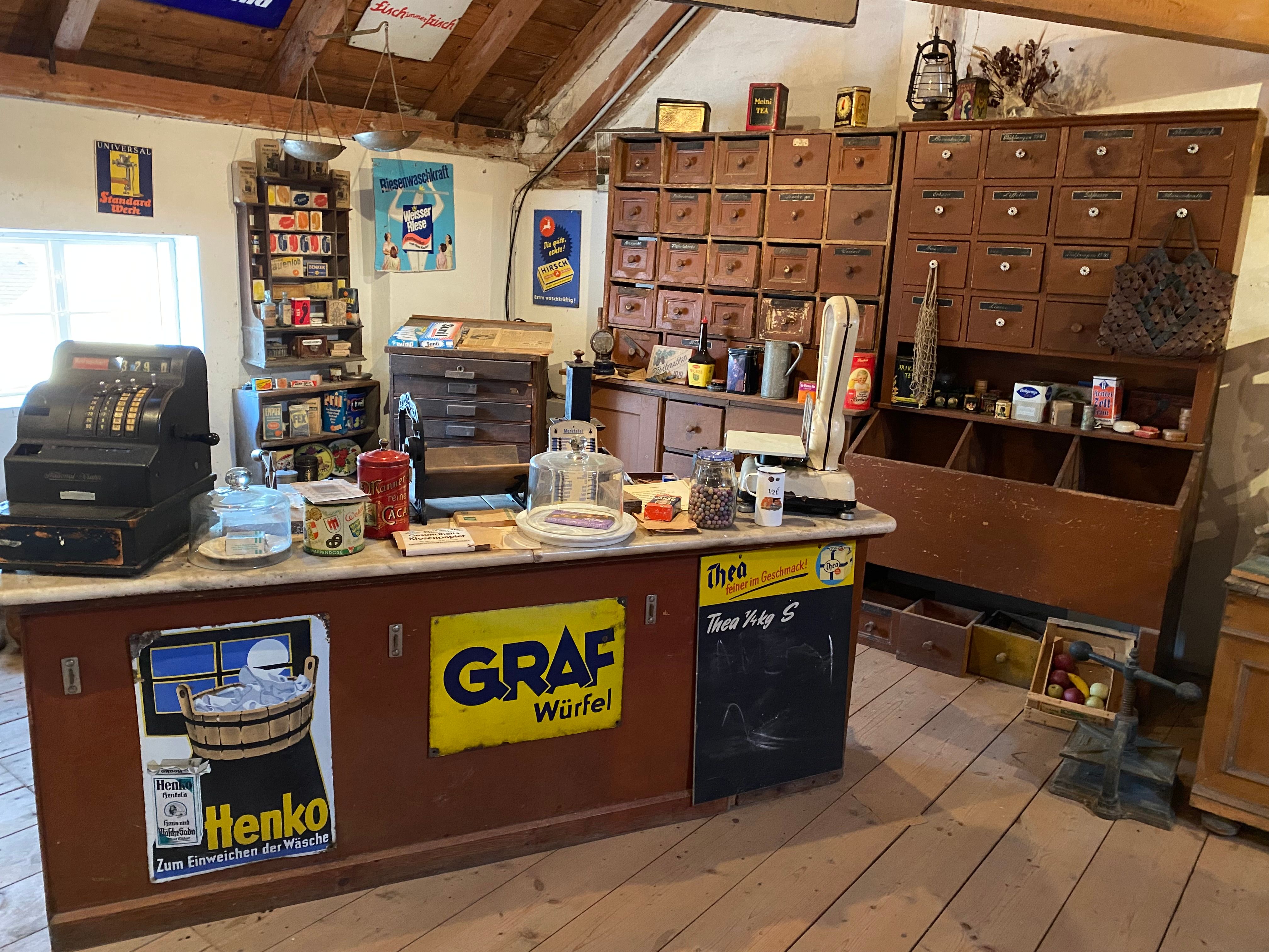 Innenansicht eines historischen Tante-Emma-Ladens mit alter Registrierkasse, Waage und Regalen voller Waren.