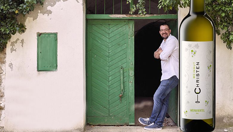 Ein Mann lehnt lässig in einer Kellertür, ins Bild gesetzt wurde eine Weinflasche.