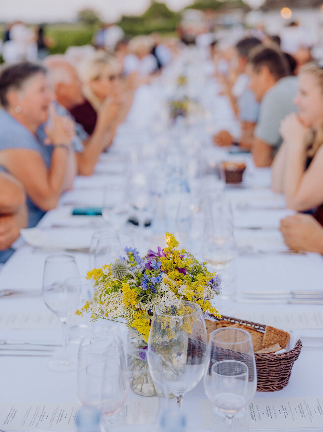 Eine lange Tafel, geschmückt mit bunten Blumen, lädt zu einem geselligen Abendessen ein. Umgeben von der malerischen Kellergasse „Alte Geringen“ genießen die Gäste die kulinarischen Köstlichkeiten der Region. Die Atmosphäre ist geprägt von fröhlichem Lachen und dem Duft von frisch zubereiteten Speisen.