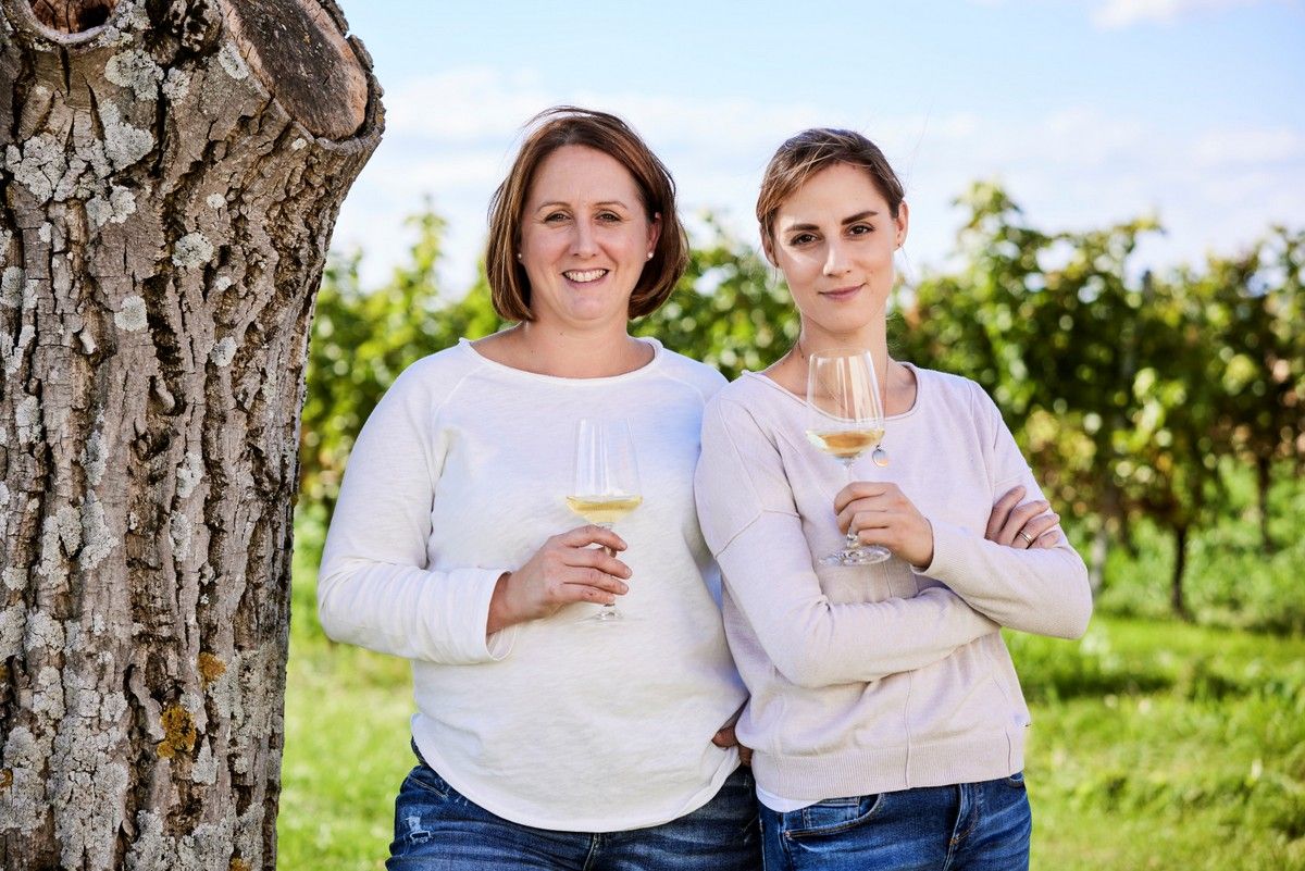 Zwei Frauen stehen mit Weingläsern vor einem Baum in einem Weinberg.