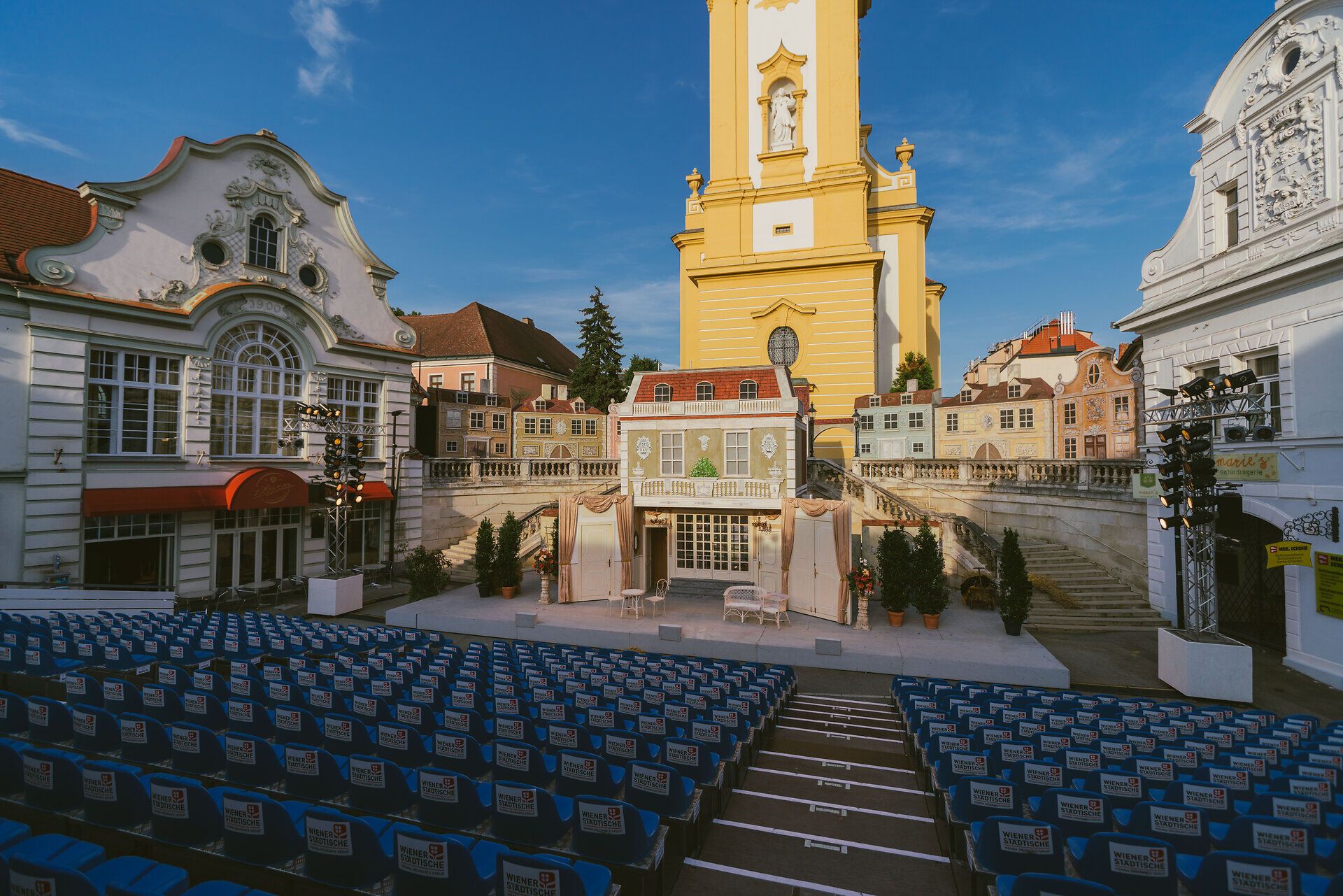 Ein malerisches Freilufttheater, umgeben von historischen Gebäuden, lädt Besucher zu unvergesslichen Aufführungen ein. Die bunten Stühle sind bereit, die Zuschauer zu empfangen, während die strahlende Sonne den Himmel erleuchtet und eine festliche Stimmung verbreitet.
