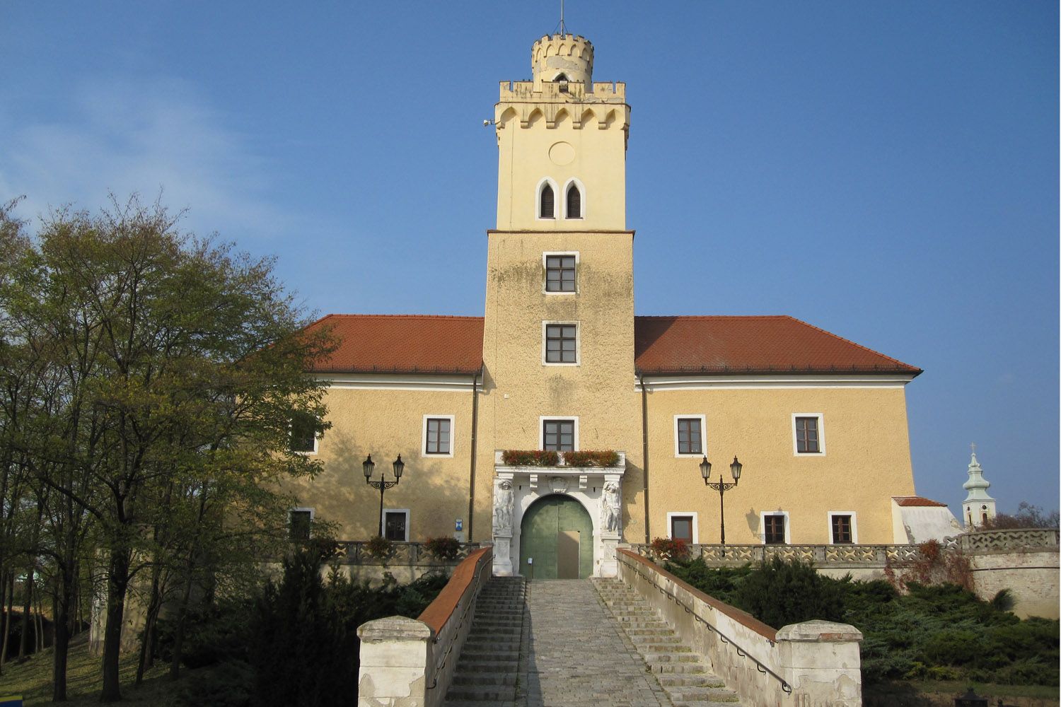 Schloss mit gelber Fassade und rotem Dach, zentraler Turm, breite Steintreppe, blauer Himmel.
