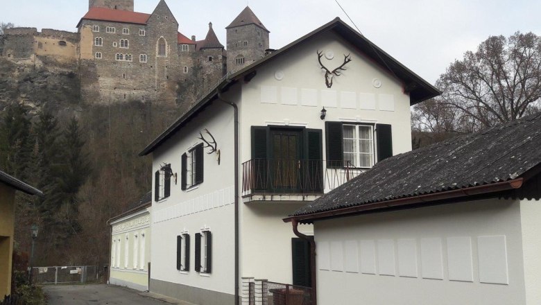 Ein weißes Haus mit grünen Fensterläden und Hirschgeweihen an der Wand, im Hintergrund eine Burg auf einem Hügel.