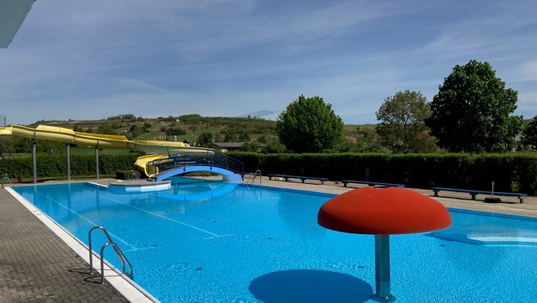 Ein Freibad mit einer gelben Wasserrutsche, einem blauen Becken und einem roten Wasserspiel in Form eines Pilzes.