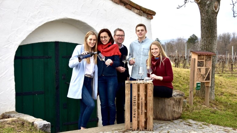 Eine Familie steht vor einem Weinkeller im Freien, mit Weingl&auml;sern und einer Weinflasche.