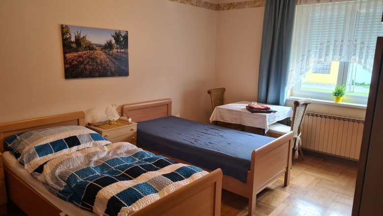 Ein einfach eingerichtetes Schlafzimmer mit zwei Einzelbetten, einem Tisch mit Stuhl und einem Fenster mit Vorhängen.