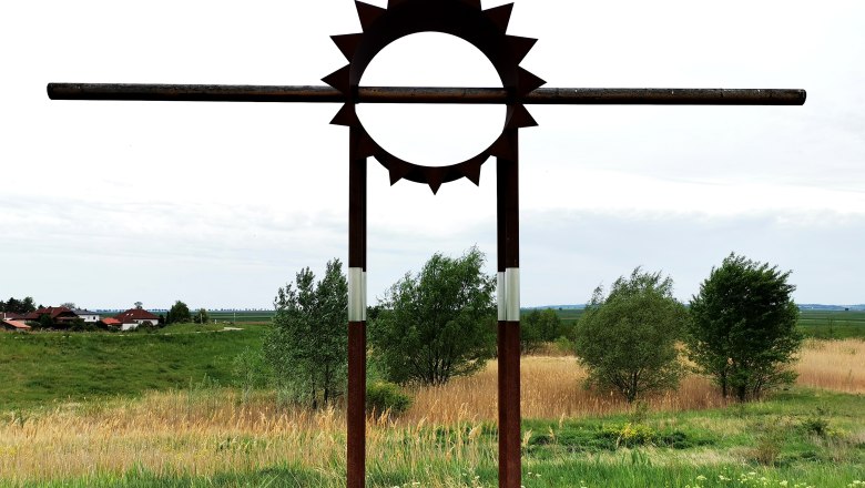 Metallskulptur in Form einer Sonne auf einer Wiese mit Bäumen im Hintergrund.