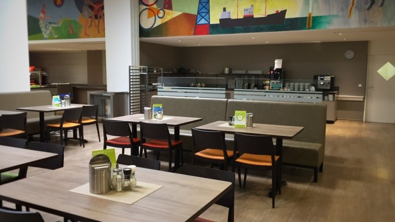 Innenansicht eines Restaurants mit leeren Tischen und Stühlen, bunte Wandmalerei im Hintergrund.