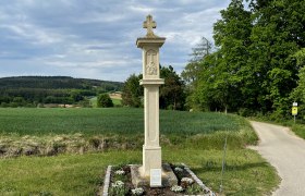 Steinkreuz auf einem Feldweg mit gr&uuml;ner Landschaft im Hintergrund.