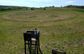Aussichtsturm bei den Kreisgräben in Glaubendorf mit Person darauf, umgeben von grüner Landschaft.