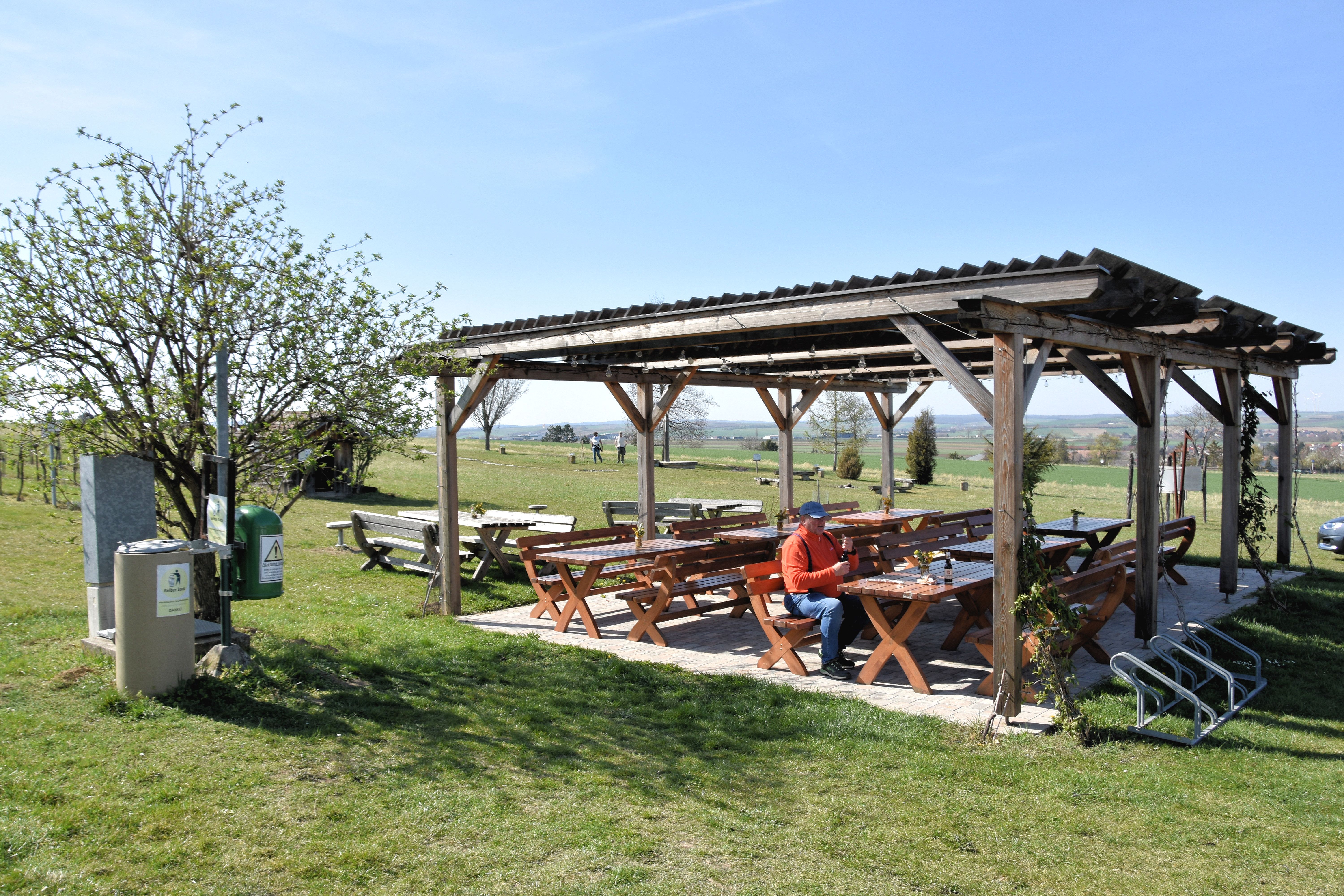 Ein überdachter Rastplatz mit Holzbänken und Tischen auf einer Wiese, eine Person sitzt dort. Im Hintergrund sind Felder und Bäume zu sehen.