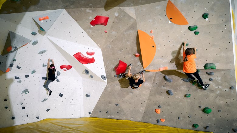 Drei Kinder klettern an einer Indoor-Boulderwand mit bunten Griffen.