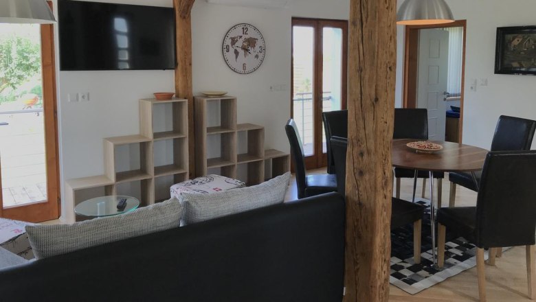 Moderner Wohnbereich mit Holzbalken, Esstisch, St&uuml;hlen, Wandregal, Uhr und Fernseher.