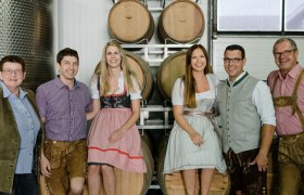 Eine Gruppe von sechs Personen in traditioneller bayerischer Kleidung steht vor Weinfässern in einem Weinkeller.