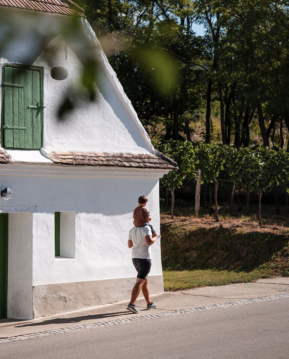 Ein Vater trägt seinen Sohn auf den Schultern und spaziert mit ihm an einem weiß gekalkten Presshaus vorbei.