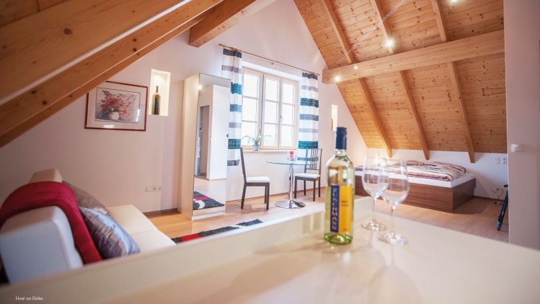 Gemütliches Dachgeschosszimmer mit Holzbalkendecke, Bett, Sofa, Tisch und Weinflasche.