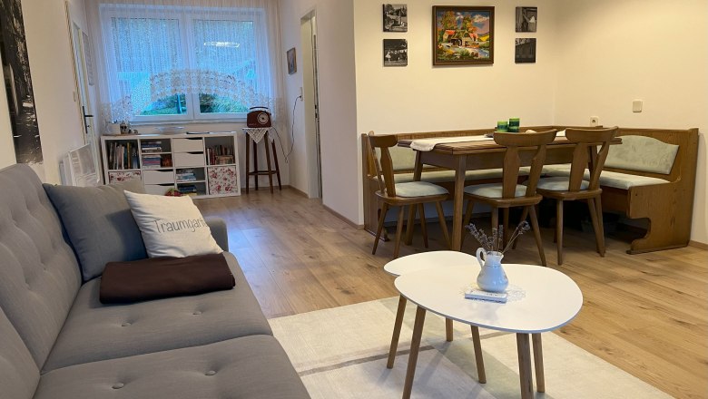 Gemütliches Wohnzimmer mit grauem Sofa, Esstisch und Stühlen, Bücherregal und Fenster mit Gardinen.