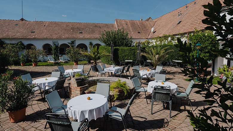 A cozy garden with round tables and chairs, surrounded by plants and an old building with a tiled roof.