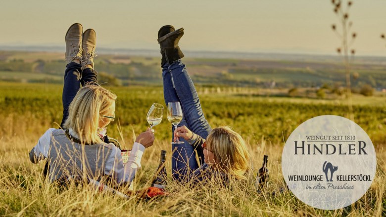 Two people are lying in a vineyard, raising their feet and holding wine glasses. The Hindler winery logo is shown.