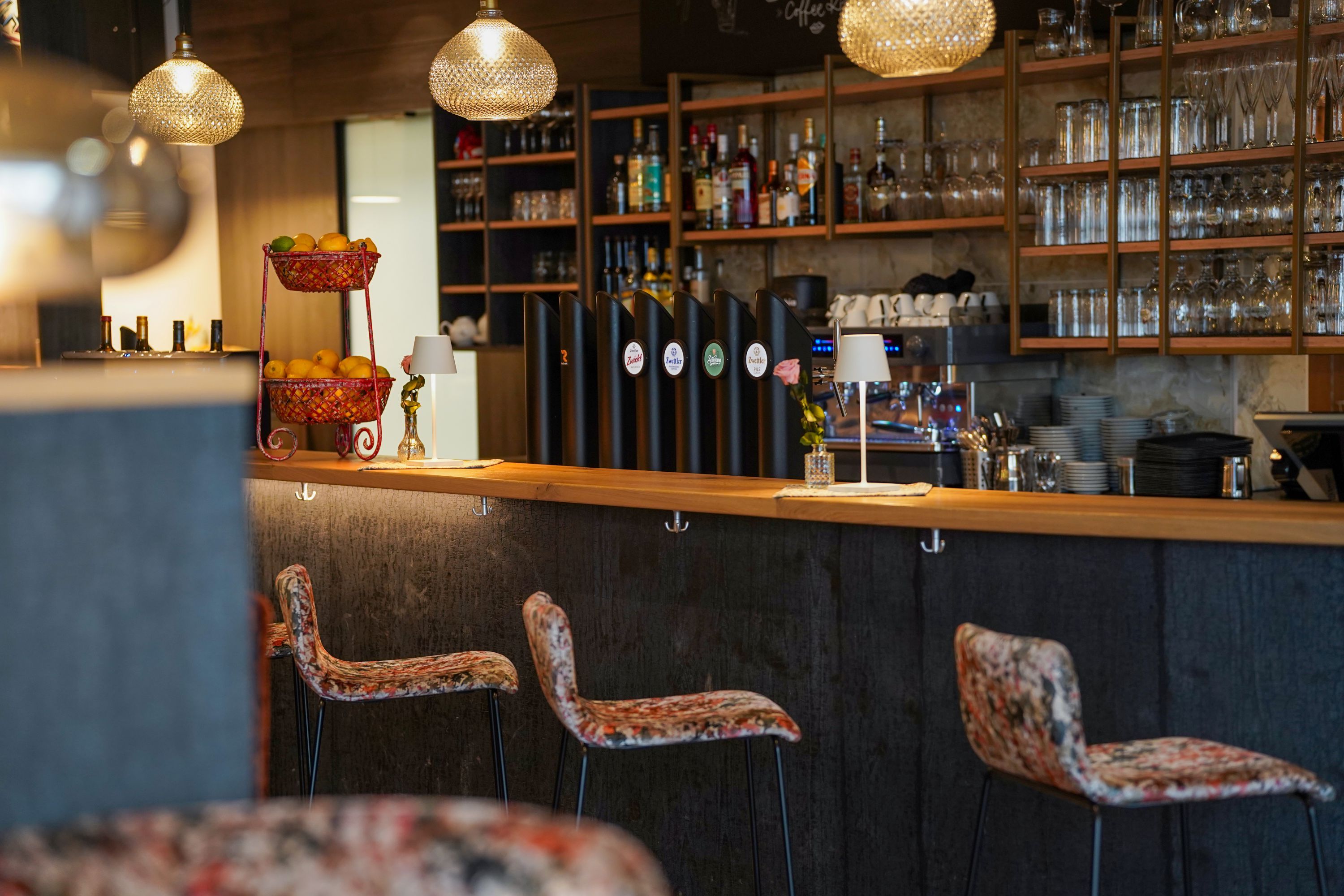 A cozy indoor bar with wooden shelves, glasses, bottles, pendant lights, and bar stools; there is a fruit basket on the counter.