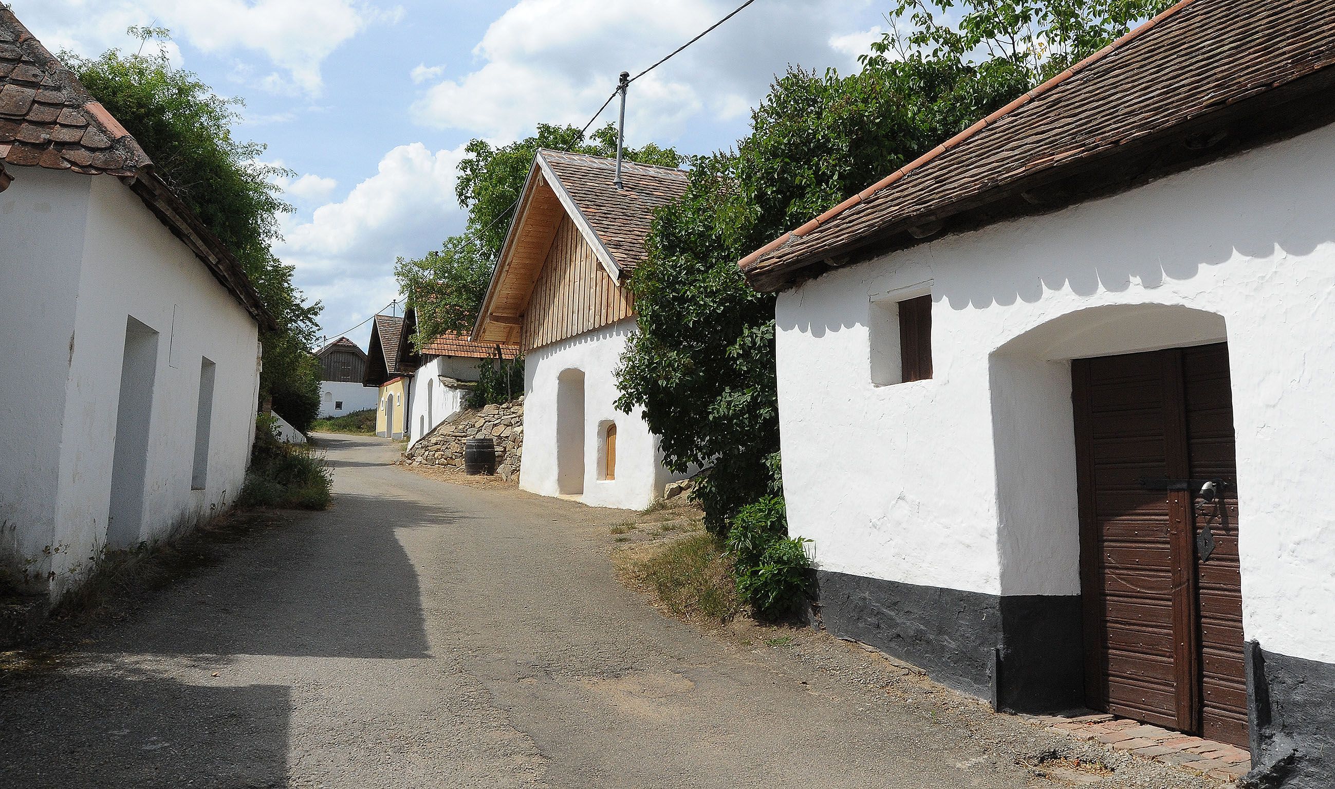 Eine malerische Gasse mit traditionellen Weinkellern in Pillersdorf, umgeben von Bäumen und blauem Himmel.