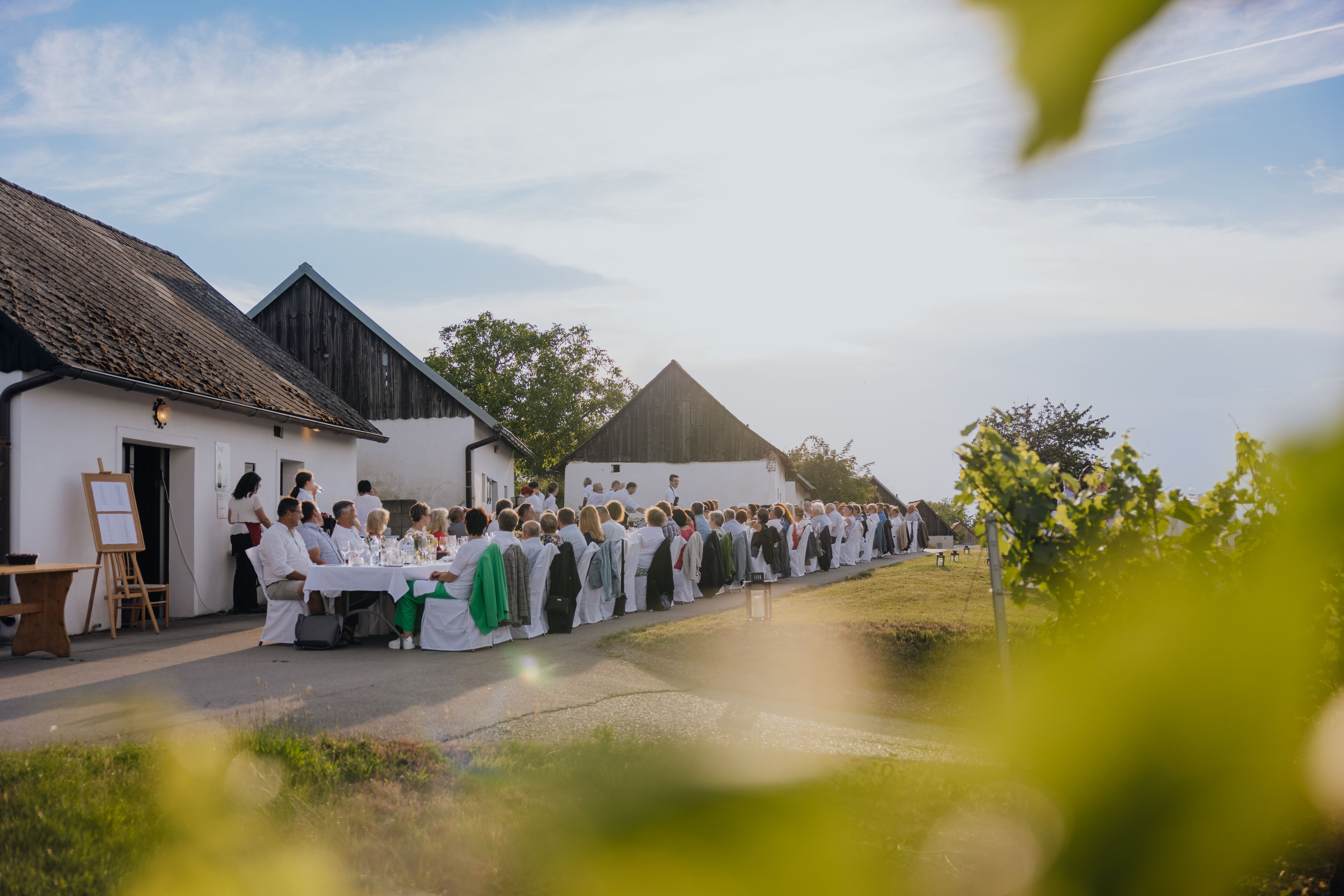 Menschen sitzen an langen Tischen im Freien vor traditionellen Gebäuden, umgeben von Natur.