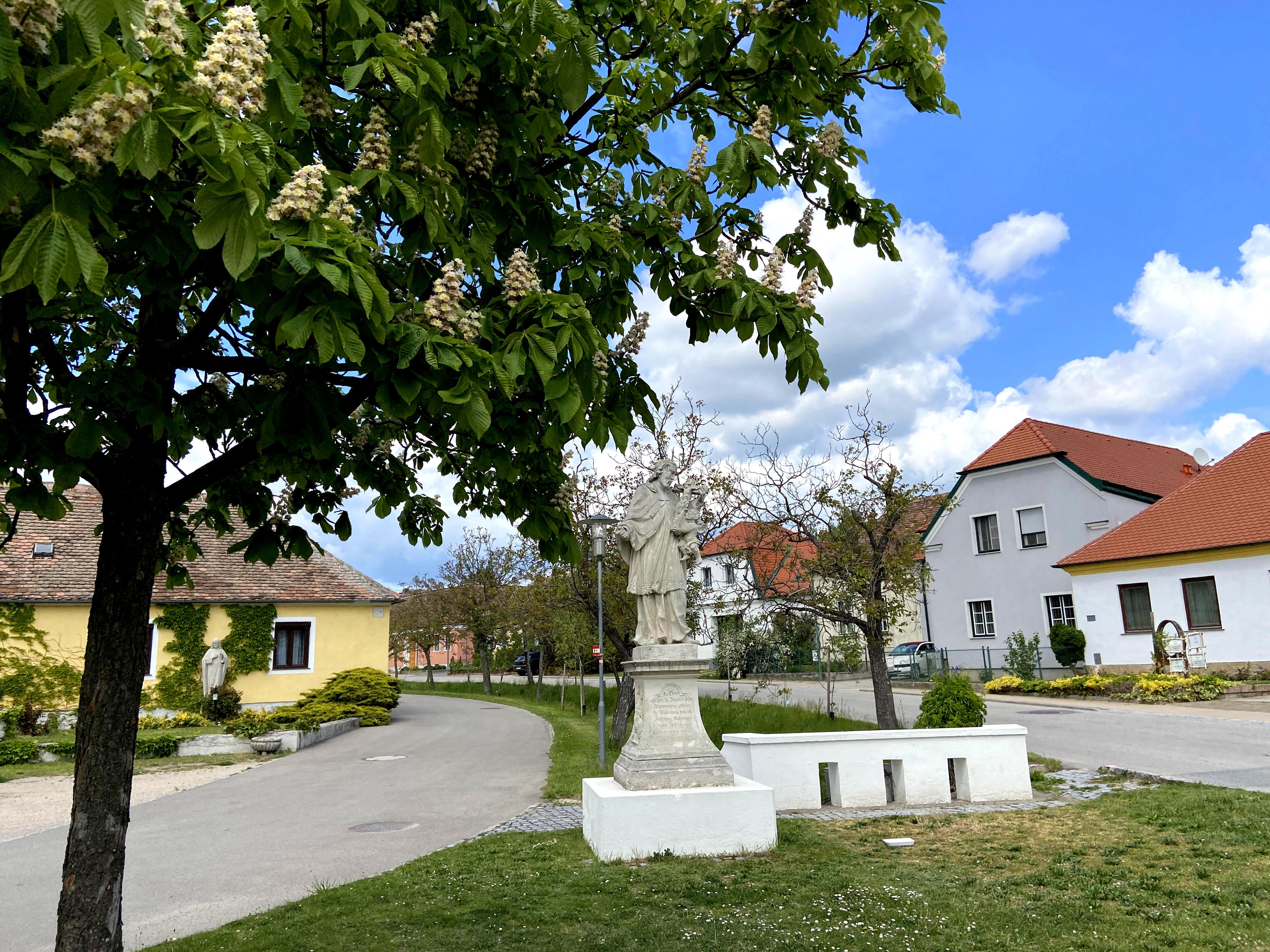 Statue des Hl. Johannes Nepomuk in Obermarkersdorf, umgeben von blühenden Bäumen und Häusern.