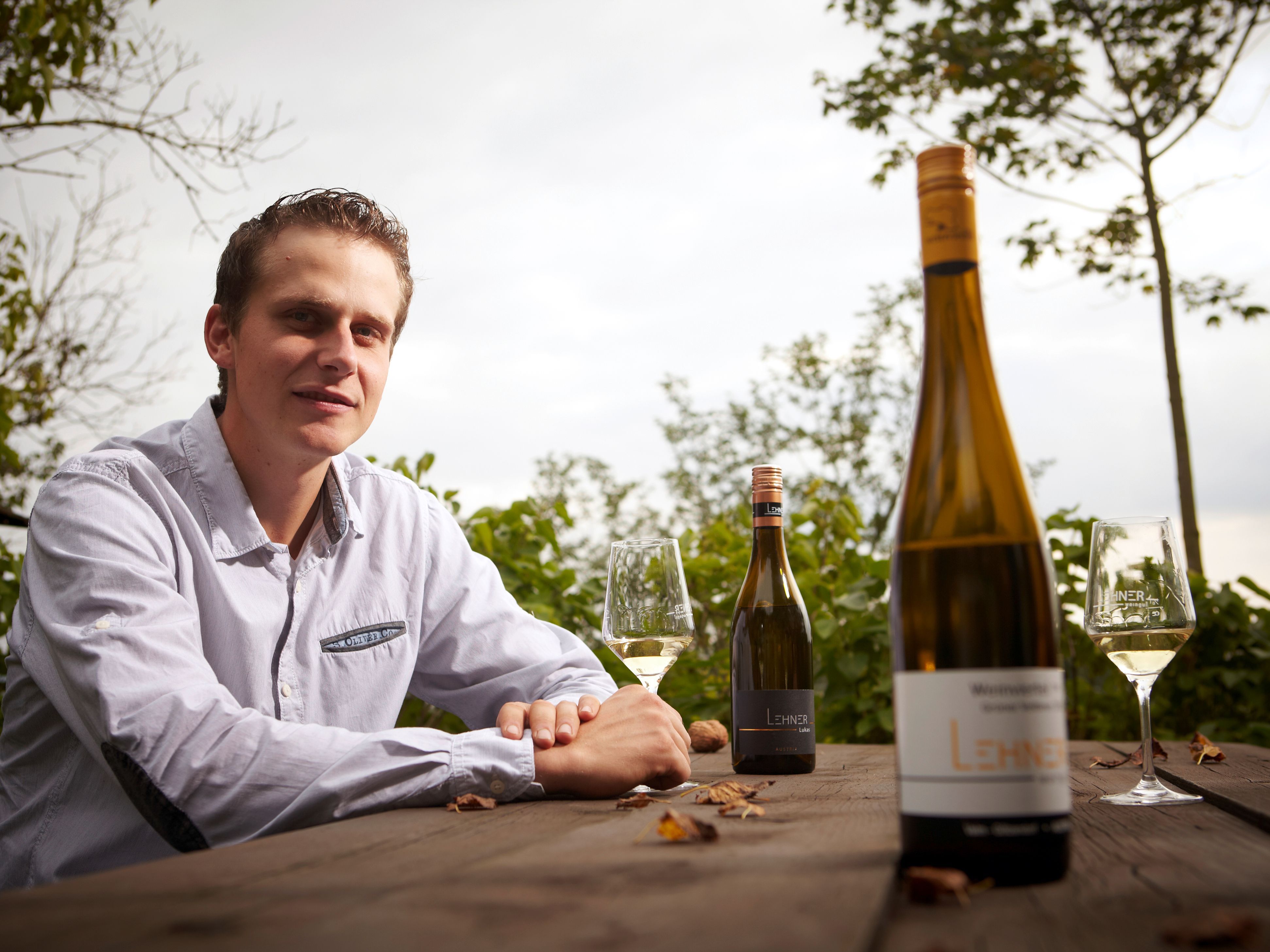 A man sits at a wooden table outside with bottles of wine and glasses.