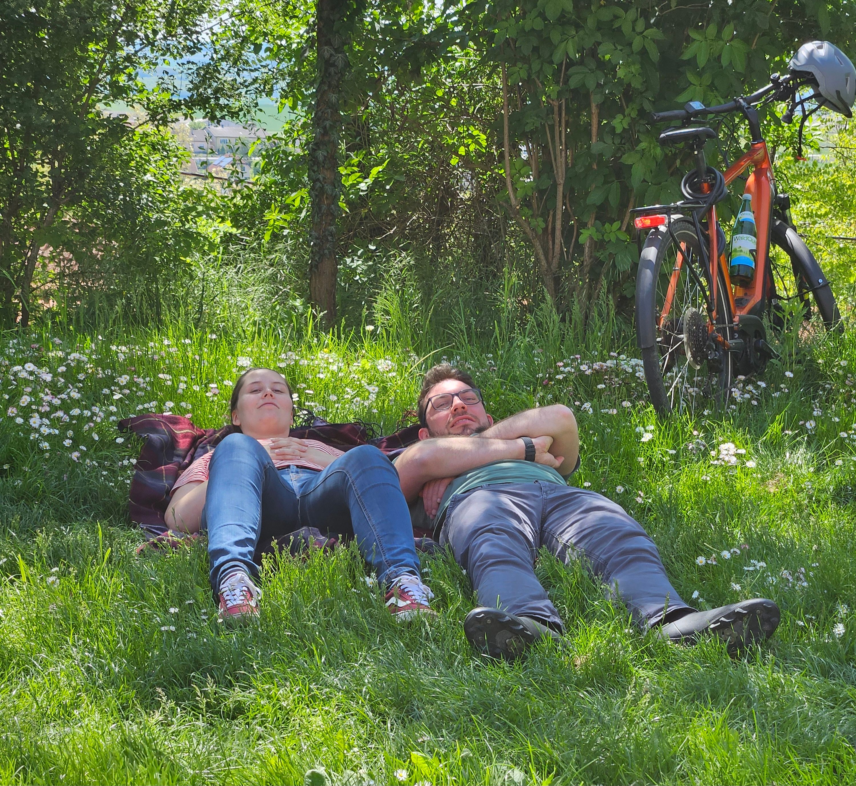 Zwei Personen liegen entspannt auf einer Wiese im Schatten, neben ihnen steht ein Fahrrad.