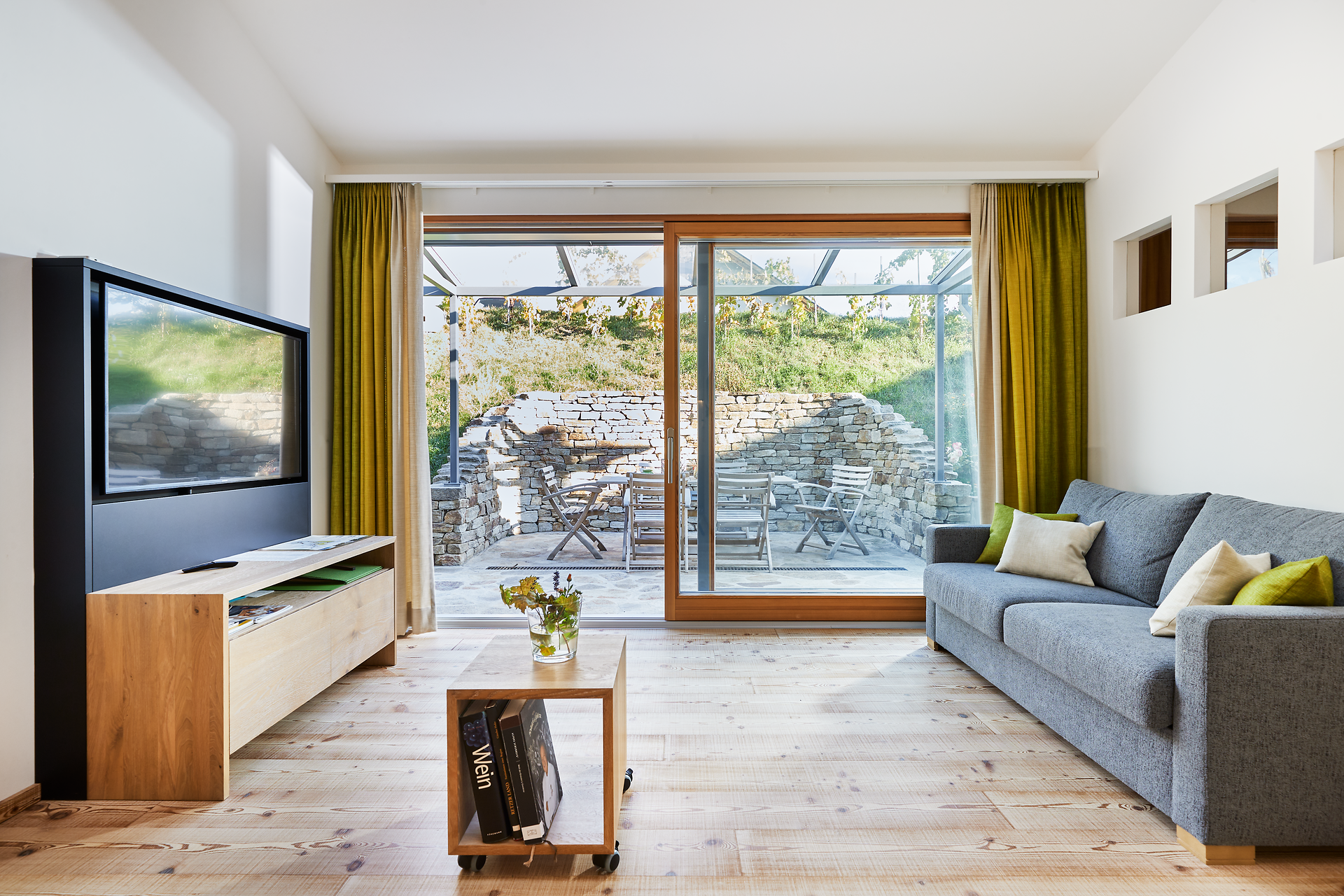 In einem einladenden Wohnraum, der mit warmen Holztönen und großen Fenstern gestaltet ist, strömt das Licht herein und schafft eine harmonische Atmosphäre. Der Blick auf die idyllische Terrasse, umgeben von sanften Hügeln und Weinreben, lädt zum Entspannen und Genießen ein. Hier wird der Aufenthalt zu einem unvergesslichen Erlebnis inmitten der Weinlandschaft.