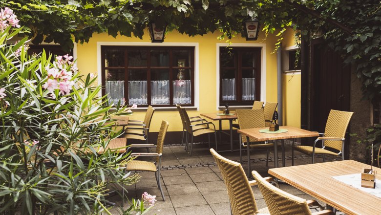 Gem&uuml;tlicher Gastgarten eines Wirtshauses mit gelber Fassade, Holztischen und Korbst&uuml;hlen, umgeben von Pflanzen und Weinreben.