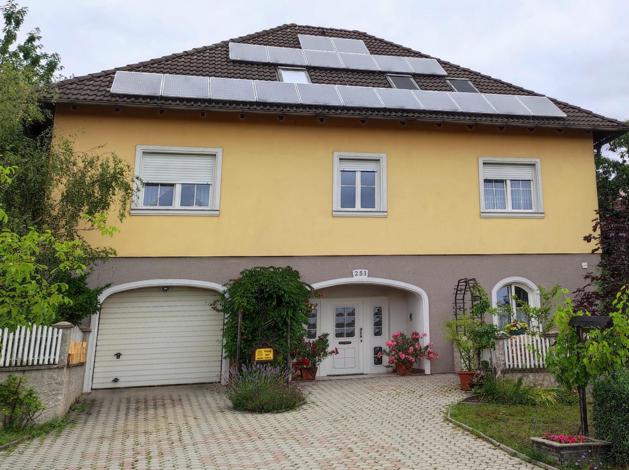 Gelbes Haus mit Solarpanels auf dem Dach, Garage und Eingang mit Blumen.