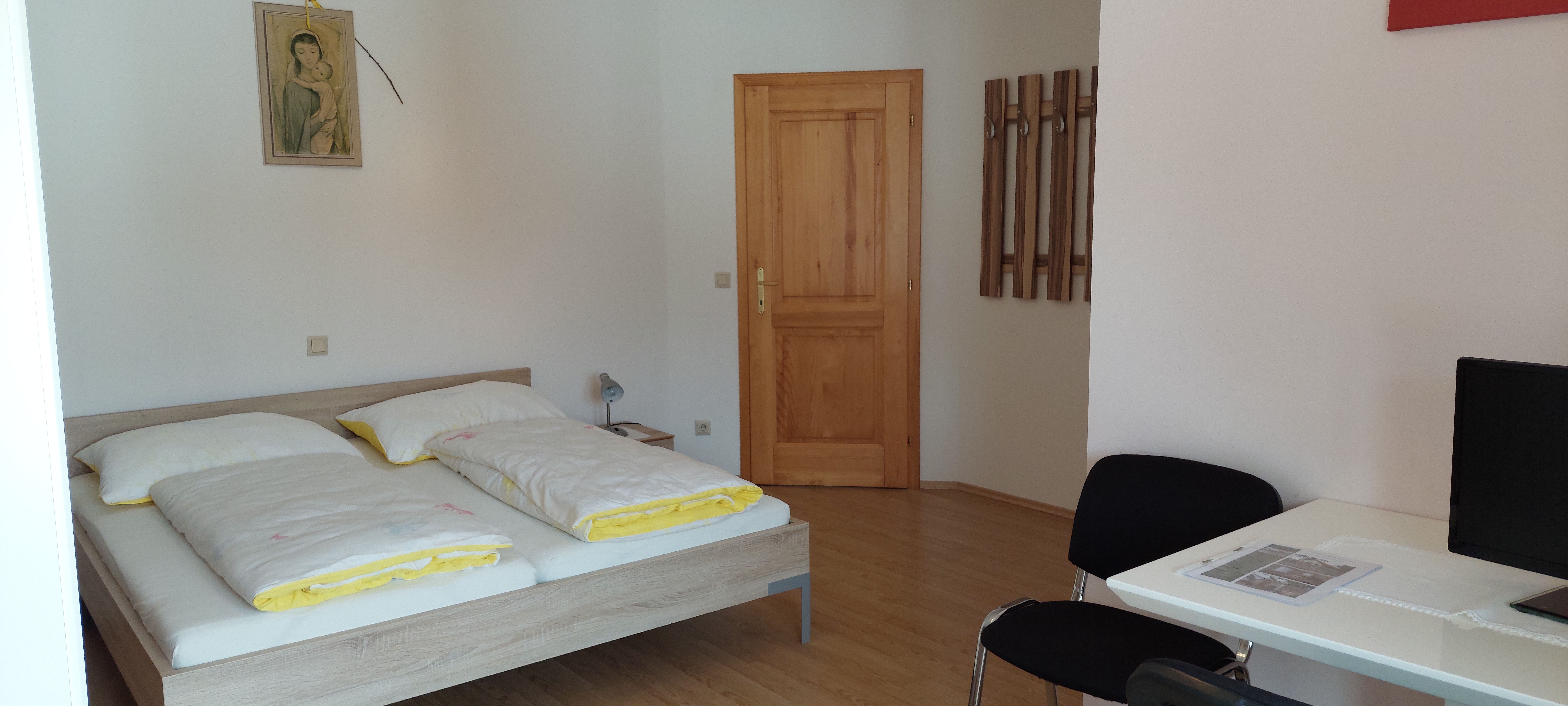 Ein einfach eingerichtetes Schlafzimmer mit Doppelbett, Holztür, Schreibtisch und Stuhl.